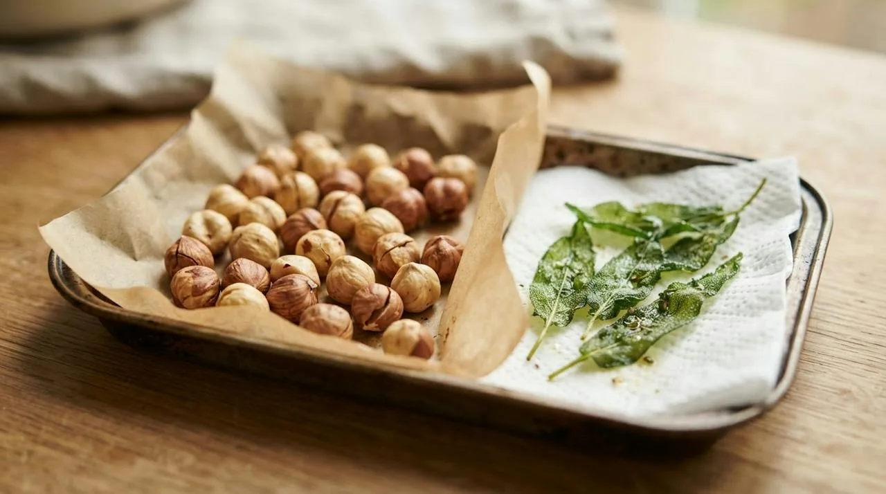 Bakplaat met licht geroosterde hazelnoten met gebarsten, lichtbruine velletjes en krokante groene salieblaadjes op keukenpapier